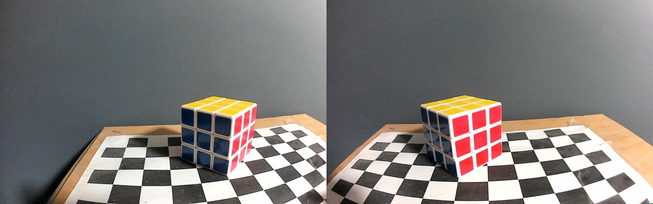 A side by side stereo image from the Mynt Eye camera depicting a rubik's cube on a black and white grid precariously taped on a cardboard box with a gray wall as the background, close up wide lens, large difference in view between the left and right cameras